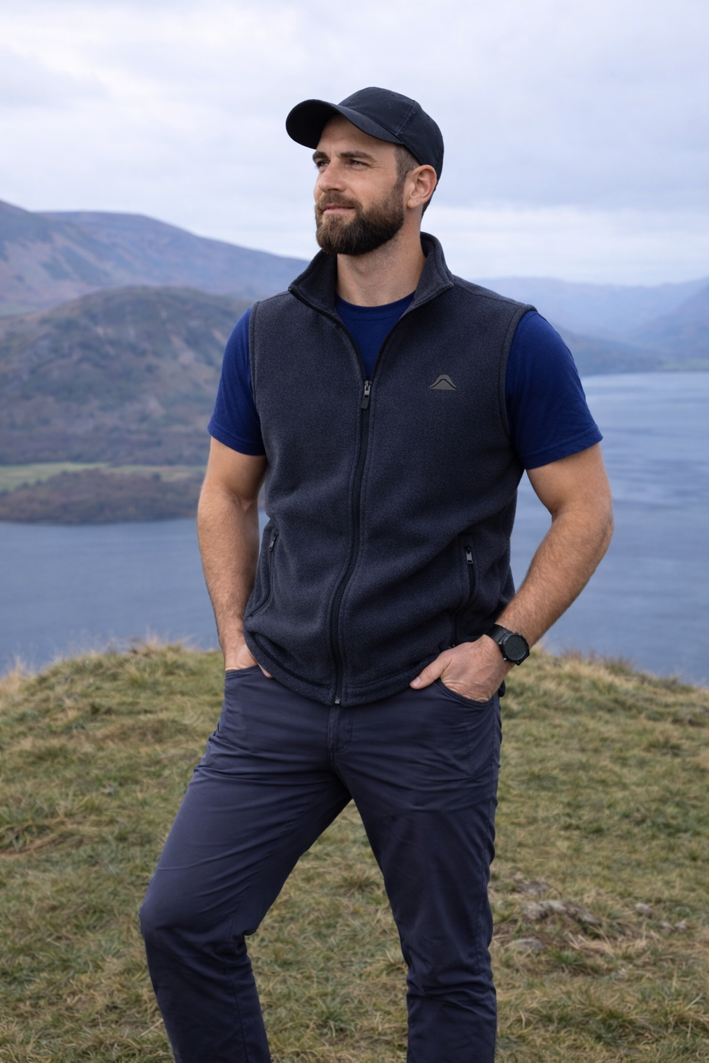 Man wearing a navy fleece vest and cap standing in front of a scenic lake with mountains.