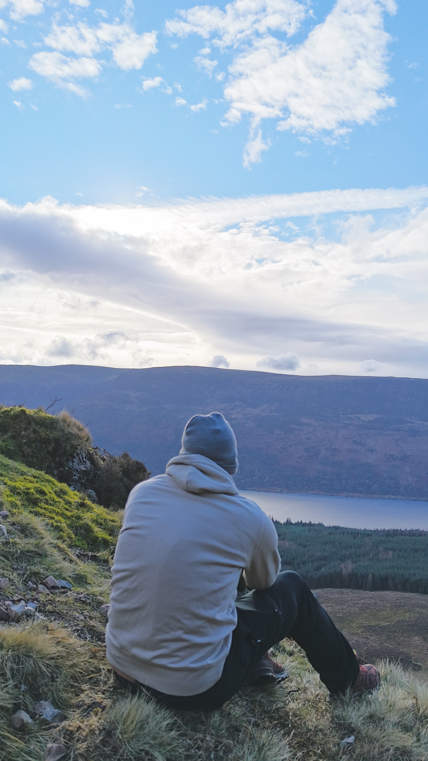 Person sitting on a grassy hill overlooking a lake with mountains in the background