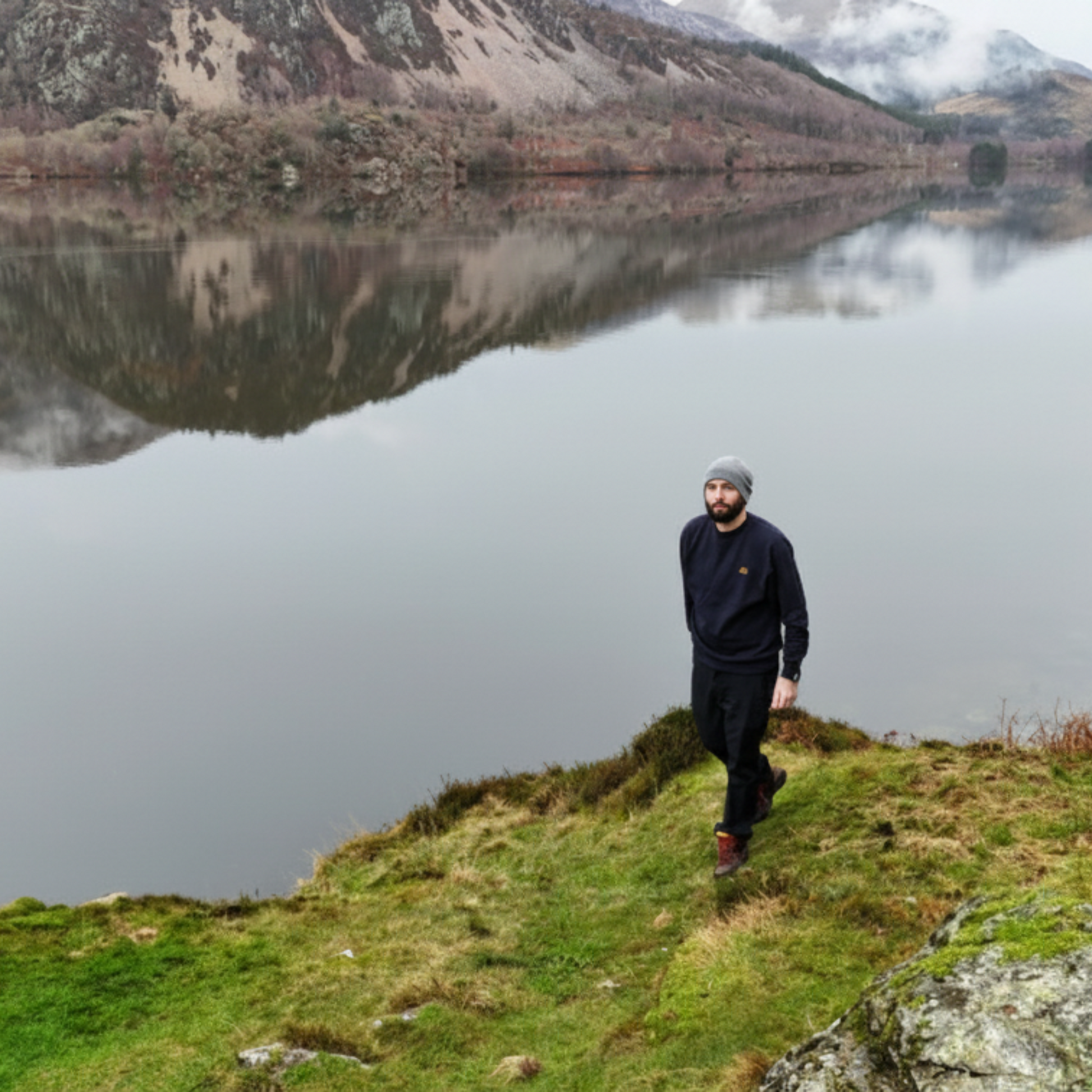 Person standing on a grassy hill overlooking a calm lake with mountains in the background